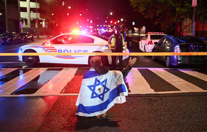 A man draped in the Israeli flag, bearing a cross and the name 'Jesus' at its center, gestures as Metropolitan Police officers secure the area outside the Capital Jewish Museum following a shooting that left two people dead in Washington, D.C., in the early hours of May 22, 2025. Two Israeli embassy staffers were shot dead late Wednesday, May 21, outside a Jewish museum in Washington by a gunman who shouted 'free, free Palestine,' authorities said, with U.S. officials and Israeli diplomats expressing shock and outrage over the killings.