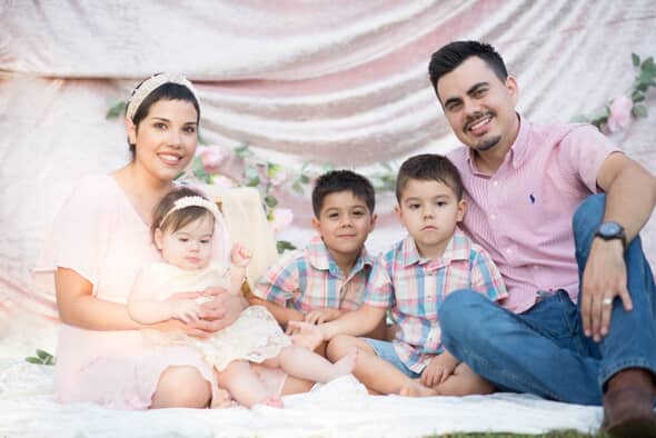 A family portrait featuring a mother holding a baby girl, with two young boys and a father sitting beside them, all smiling in front of a softly draped backdrop adorned with floral decorations.