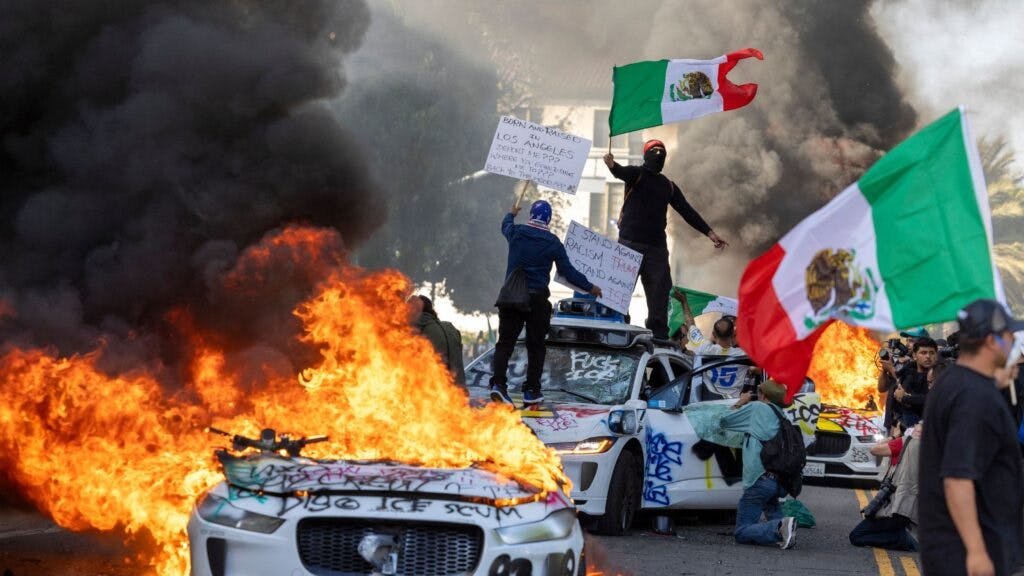 Demonstrators wave flags from atop a wrecked car, standing beside burning Waymo vehicles, as protesters clash with law enforcement in the streets surrounding the federal building during a protest following federal immigration operations in Los Angeles, California, on June 8, 2025.