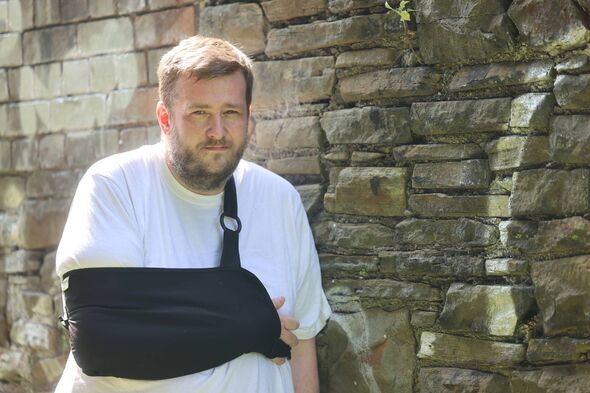 Michael Gwyther, with a stern expression on his face, right arm in a black sling, standing against a stone wall