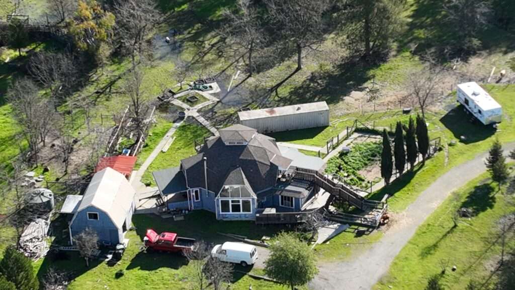 From above, a geodesic dome home with a couple buildings nearby, along with a garden, a camping-style trailer, two vehicles, and green grass and trees.