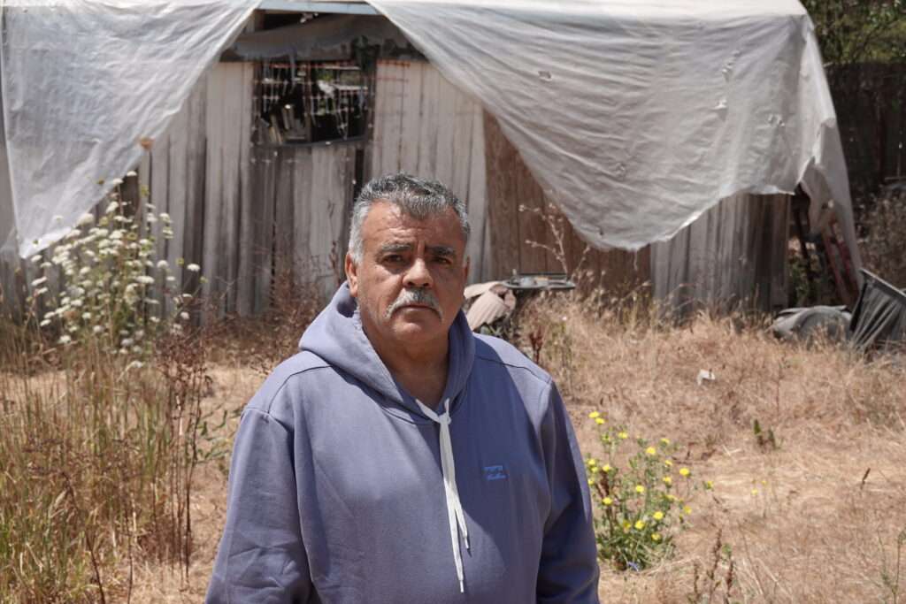 A man in a gray hoodie, with gray hair and a mustache, stands in front of tall brown grass and other plants in front of a structure covered by a white plastic tarp.