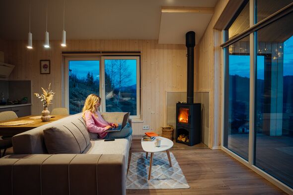 Young woman works on laptop in modern living room in mountain home with fire burning in cast iron fireplace