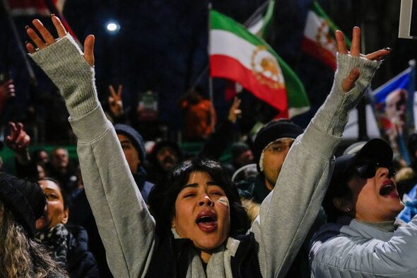 A group of individuals, many of whom are holding their arms aloft, gather in a public space. Some are waving flags, and the atmo