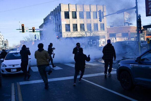 A group of individuals is seen running across a street amidst smoke, with a building and traffic signals visible in the backgrou