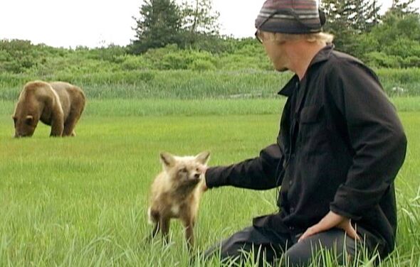 An individual dressed in a black jacket and a hat is seated in a grassy field, gently holding a small, light-colored animal. In 