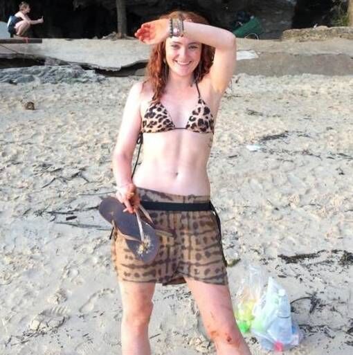 An individual is standing on a gravel beach, dressed in a patterned bikini, holding a surfboard in one hand and touching their h