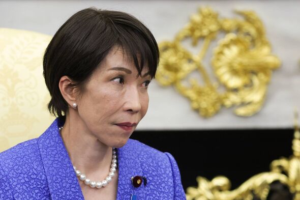 An individual in formal attire, adorned with a pearl necklace and a microphone, stands in front of an ornate backdrop featuring 