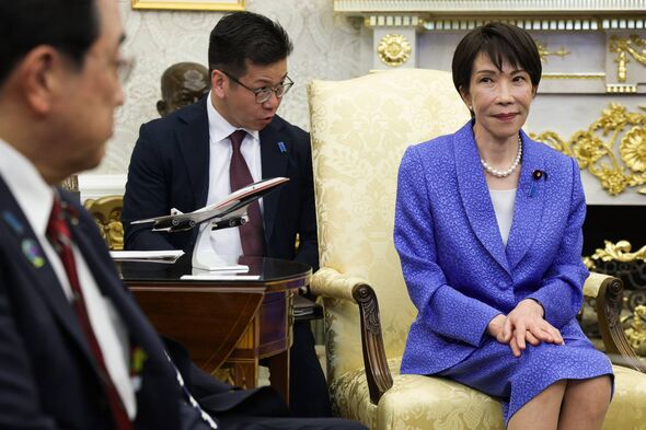 A formal meeting is taking place in an elegant room. A person in a blue dress is seated on a chair, while two individuals dresse
