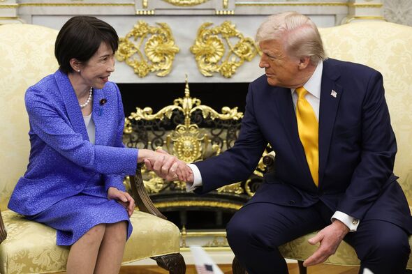 An individual in formal attire, seated, engages in a handshake with another individual, also in formal attire, in an opulent set