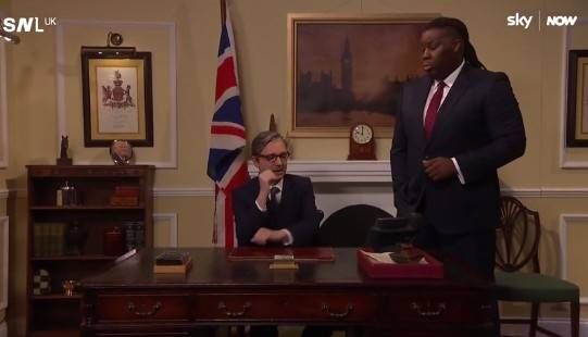 Two individuals are present in a formal office setting. One person is seated at a desk, while the other stands nearby. The room 