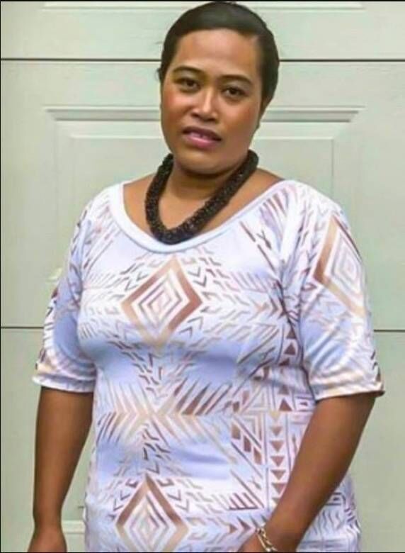 An adult female individual dressed in a white shirt adorned with floral patterns and a black necklace stands in front of a plain An adult female individual dressed in a white shirt adorned with floral patterns and a black necklace stands in front of a plain