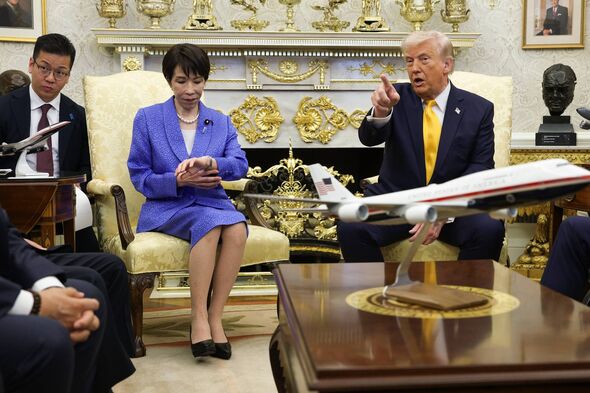 In an official meeting, a group of individuals are seated around a wooden desk in a formal setting. A male figure is prominently