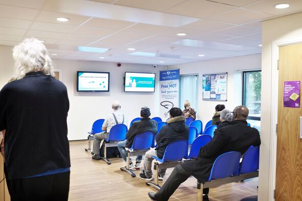 Person standing at reception while a busy waiting room in a health centre is busy and full with peop Person standing at reception while a busy waiting room in a health centre is busy and full with peop