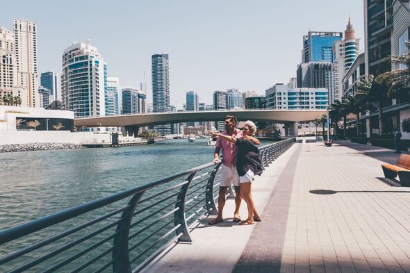 Couple walking along a waterfront promenade with high-rise skyscrapers and a bridge in the background.Urban tourism, modern travel and love concept.