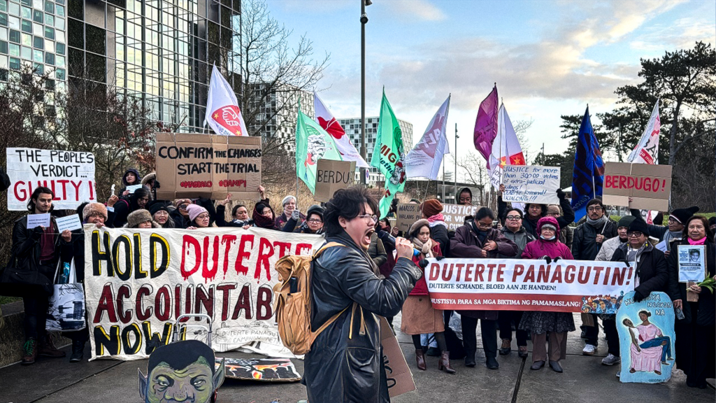 A man in a black leather jack is yelling into a microphone while wearing an orange-yellow backpack, and his fellow anti-Duterte protestors are holding signs and waving flags.