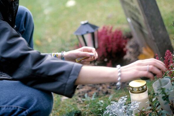 An individual is seen kneeling and placing a candle on a memorial site, surrounded by a variety of greenery and flowers, with a 