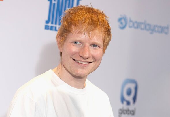 An individual with red hair is standing in front of a wall adorned with logos, wearing a white t-shirt and smiling at the camera