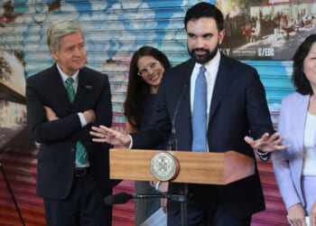 New York Mayor Zohran Mamdani stands at a little podium with his arms wide in front of a grafitti-art filled wall. Three associates surround him.