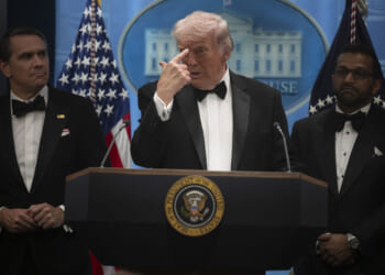 Donald Trump in a tux points to his forehead during a press briefing. He's flanked by Kash Patel and Todd Blanche.