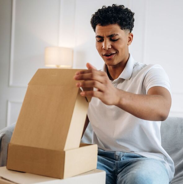 Young male unboxes a cardboard package while sitting on a sofa in a modern living room with a lamp and neutral-colored walls in the background